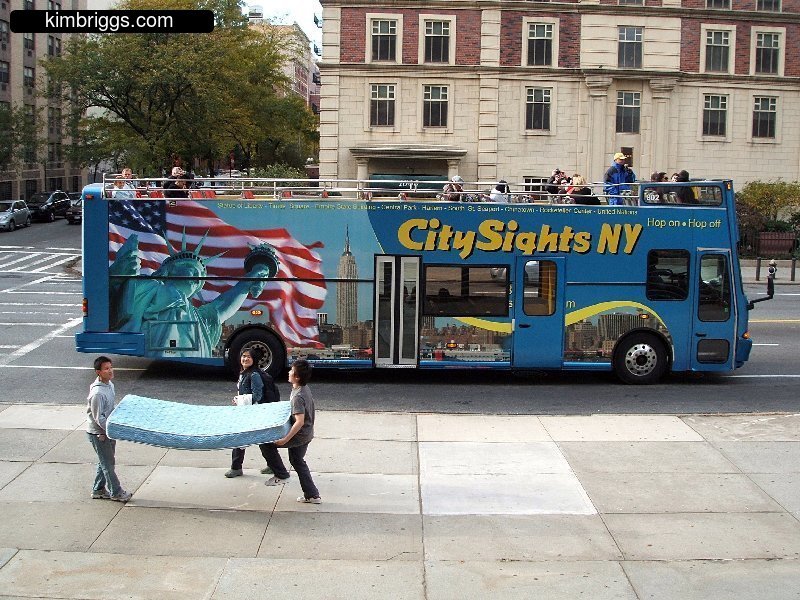 People carrying mattress near City Sights bus, NYC.